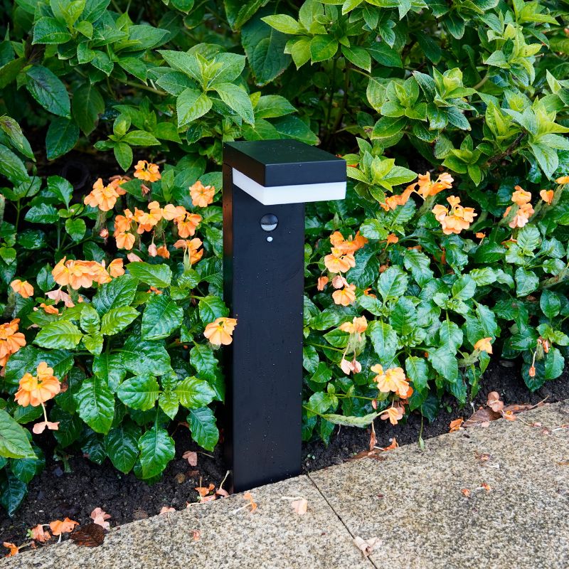 This is a modern, sleek black outdoor floor lamp with a motion sensor, placed amidst lush greenery and blooming orange flowers, providing elegant lighting for garden spaces.