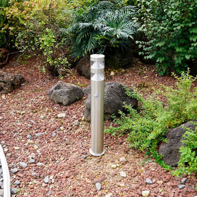 Silver outdoor floor lamp with motion sensor, designed with a sleek cylindrical shape, standing in a garden with rocks and plants around it.
