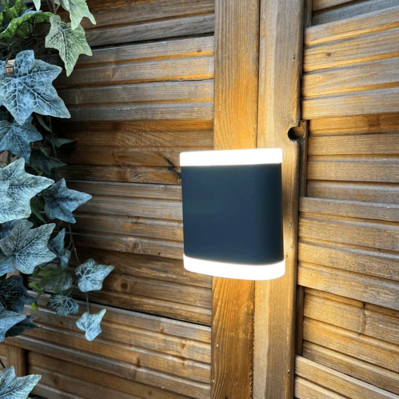 A black outdoor wall lamp with a sleek design, mounted on a wooden fence and surrounded by ivy, offering a stylish and functional lighting solution.